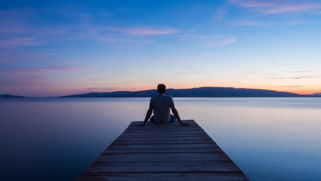 Man sitting on dock looking out over water at sunrise, representing mindfulness, the practice of reframing, which leads to clarity, direction, and empowerment.